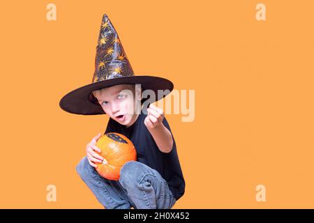 Halloween Kinder. Kleiner emotionaler Junge in einem Zauberer-Hut, der einen orangefarbenen Kürbis in den Händen hält und auf einem orangefarbenen Hintergrund eine Süßigkeit zeigt. Das Kind will Stockfoto