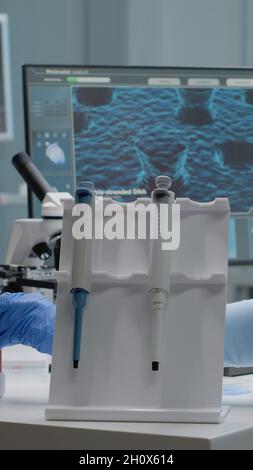Wissenschaftlerin sitzt im Labor mit chemischer Technologie auf dem Schreibtisch. Biochemie Arzt mit Pipette, Blutvacutainser, Mikroskop, Eingabe am Computer für mikrobiologische Experiment Stockfoto