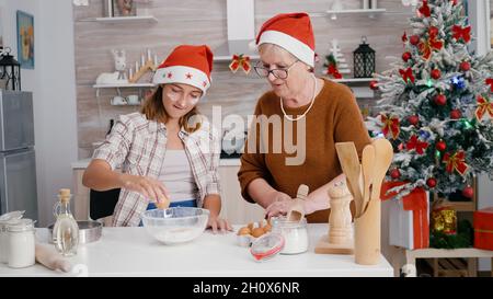 Enkelin mit Enkeltochter, die in einer Küchenschüssel Eier bricht, mit Mehlzutat, um hausgemachten Teig zuzubereiten. Glückliche Familie genießen die Herstellung von Weihnachtskeks köstlichen Dessert während der Weihnachtszeit Stockfoto