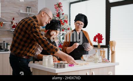 Großeltern lehren Enkelin, wie man Weihnachten hausgemachte Lebkuchen mit Cookies Form in dekorierten kulinarischen Küche zu machen. Glückliche Familie verbringt Zeit zusammen während der weihnachtsferien Stockfoto