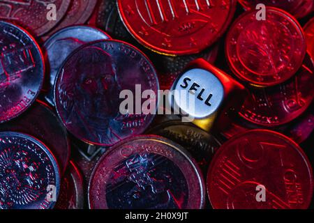 Ein Metall würfelt in einem Haufen Münzen aus verschiedenen Ländern unter einem roten Licht. Am Rand der Würfel befindet sich das Wort „verkaufen“. Stockfoto