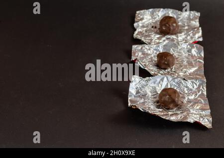 Runde Pralinen in ungewickelter Silberverpackung auf schwarzem Hintergrund. Zerknitterte Folie mit drei Schokoladen-Leckereien. Selektiver Fokus. Stockfoto