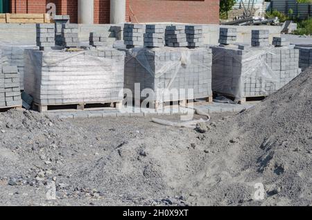 Bauindustrie, Baustoffe. Paletten aus grauen Pflasterfliesen und einem Sandhaufen. Neue Zementblöcke und Bordsteine in Plastikfolie verpackt. Keine pe Stockfoto