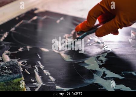 Kochfeld mit weißem Reiniger. Hand in orange Handschuh mit Metallschaber und Schwamm. Selektiver Fokus. Elektroherd Reinigung Hausreinigung Konzept Stockfoto