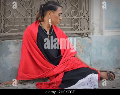 Der hübsche junge, nicht binäre Latino trägt einen roten mexikanischen rebozo und setzt sich auf den Bürgersteig vor einem traditionellen schmiedeeisernen Fenstergrill. Stockfoto