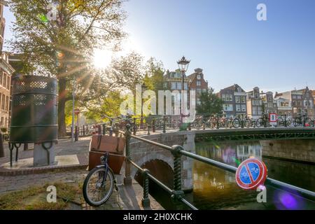 Niederlande. Sonniger Morgen in Amsterdam. Traditionelle Häuser am Kanal. Viele Fahrräder in der Nähe des Zauns der Brücke. Street Male Toilette Stockfoto