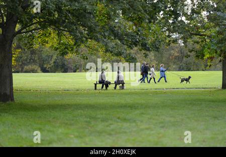 London, England, Großbritannien. Regent's Park: Zwei Frauen reden auf einer Bank, Menschen gehen mit einem Hund vorbei Stockfoto