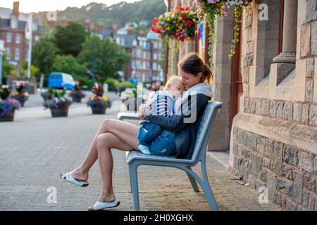 Mutter, die auf einer Bank im Zentrum des wunderschönen alten Dorfes sitzt und ihren kleinen Jungen hält, lächelt und umarmt Stockfoto