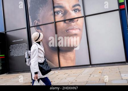 Eine farbige Frau geht unter der großen Plakatwand, ein Porträt des englischen Fußballspielers Marcus Rashford, außerhalb der Strand-Filiale der Coutts Bank, am 14. Oktober 2021 in Westminster, London, England. Marcus Rashford wurde kürzlich von der University of Manchester mit einem Ehrendoktortitel in Anerkennung seiner politischen Kampagne für die benachteiligten (insbesondere Schulmahlzeiten) und seine Philanthropie ausgezeichnet. Er spielt derzeit für Manchester United und ist in der englischen Nationalmannschaft. Er wurde auch Opfer von rassischem Missbrauch im Internet. Stockfoto