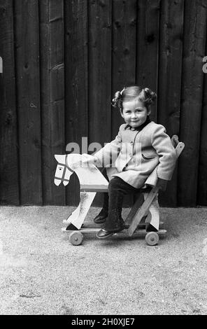 1960er Jahre, historisch, draußen auf einem flachen Kiesgebiet, ein kleines Mädchen im Mantel, das auf ihrem Reitspielzeug sitzt, ein Holzstoß auf Rädern, England, Großbritannien. Stockfoto