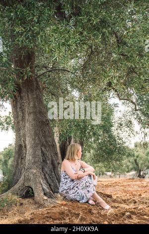 Blonde Haare Mädchen sitzt in der Nähe eines schönen weltlichen Olivenbaum in einem landwirtschaftlichen Feld ähnlich einer Skulptur im Herbst, vertikal Stockfoto