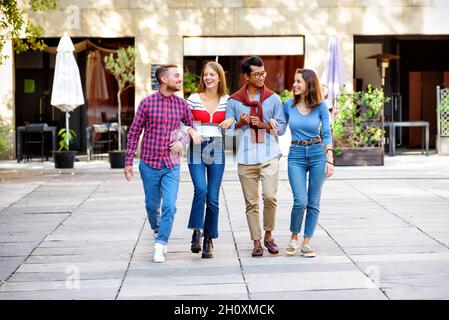 Die ganze Länge der fröhlichen, vielfältigen Freunde in legerer Kleidung hält die Hände und geht auf der Straße gegen Gebäude in der Stadt an sonnigen Tagen Stockfoto