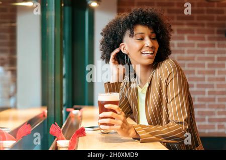 Seitenansicht einer fröhlichen jungen afroamerikanischen Frau mit stilvollem Afro-Haar in gestreiftem Blazer und einem Glas kaltem Bier, während sie sich auf der Theke ausruhte Stockfoto