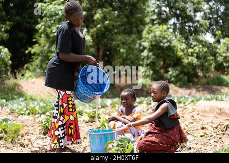 Beunruhigte junge schwarze afrikanische Frauen, die Wasser in einen Eimer am Dorfbrunnen gießen, wobei zwei kleine Mädchen darauf warten, dass der Behälter gefüllt wird Stockfoto