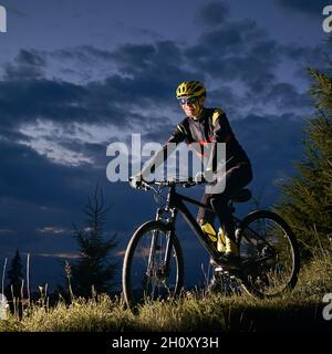 Lächelnder junger Mann, der mit dem Fahrrad bergab fährt, mit dem wunderschönen blauen Abendhimmel im Hintergrund. Männlicher Radfahrer, der nachts den grasbewachsenen Hügel hinunterradelt. Konzept von Sport, Radfahren und aktiver Freizeit. Stockfoto