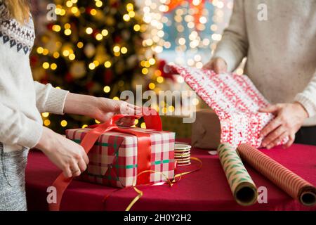 Mann und Frau bereiten sich auf Weihnachten vor. Pairle in warme Pullover Geschenkverpackung. Geschmückter Weihnachtsbaum, Schachteln mit Geschenken, buntes Geschenkpapier Stockfoto