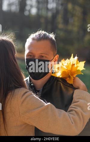 Gesundheitswesen, Coronavirus, Freizeit, Beziehungskonzept. Mann mit schwarzer Medizin Gesichtsmaske umarmt Frau im Park während sonnigen und kalten Herbsttag. Stockfoto