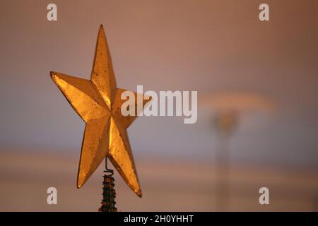 Nahaufnahme eines riesigen goldenen Sterns als Weihnachtsbaum-Topper mit einigen oberen Ästen des Baumes. Warmes und gemütliches Licht. Fröhliches und fröhliches Farbbild. Stockfoto