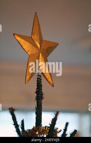 Nahaufnahme eines riesigen goldenen Sterns als Weihnachtsbaum-Topper mit einigen oberen Ästen des Baumes. Warmes und gemütliches Licht. Fröhliches und fröhliches Farbbild. Stockfoto