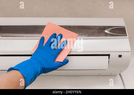 Die Hand eines Mannes in einem blauen Gummi-Haushaltshandschuh wischt und reinigt die Klimaanlage. Wartung und Reinigung von Innenräumen. Stockfoto