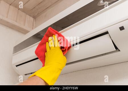 Die Hand eines Mannes in einem roten Haushaltshandschuh wischt und reinigt die Klimaanlage. Wartung und Reinigung von Innenräumen. Stockfoto