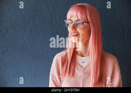Schöne Frau mit rosa Haaren und Brille trägt Pullover steht in der Nähe der blauen Wand Stockfoto