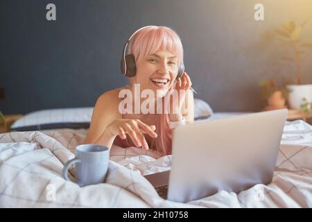 Fröhliche Frau mit Kopfhörern hört Musik auf dem Laptop auf dem Bett Stockfoto