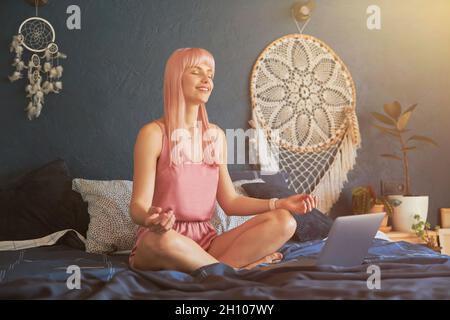 Das hübsche lächelnde weibliche Model meditiert neben dem Laptop auf einem großen Bett an der dunkelblauen Wand Stockfoto