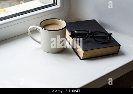 Weiße Tasse mit Kaffee, Buch und Gläsern liegen auf der Fensterbank. Bildungs- und Entspannungskonzept Stockfoto