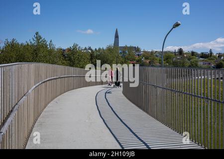 REYKJAVIK, ISLAND - 12. Juni 2021: Menschen, die über die Njardargata-Überführung zur Hringbraut Road gehen. Hallgrimskirkja-Kirche im Hintergrund. Sonniger Tag. Stockfoto