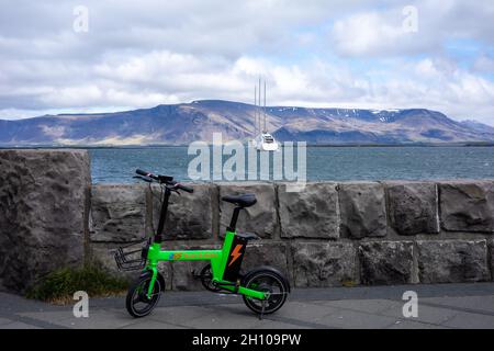 REYKJAVIK, ISLAND - 11. Juni 2021: Grünes E-Bike am Ufer von Reykjavik. Eine Luxusyacht, die in der Bucht vor Anker liegt, mit Blick auf den Berg Esja. Stockfoto