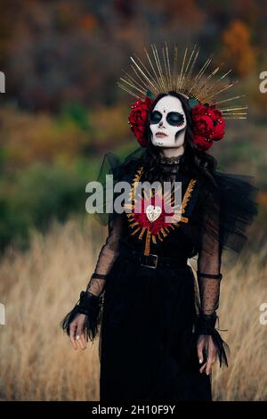 Junge Frau mit Zuckerschädel Make-up und roten Rosen in schwarzem Kostüm des Todes als Santa Muerte ist vor dem Hintergrund des Herbstwaldes gekleidet. Tag des Stockfoto