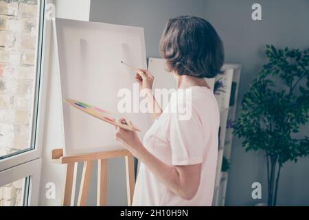 Foto von niedlichen süßen alten Frau gekleidet casual Outfit Zeichnung Bild halten Palette Pinsel drinnen Wohnzimmer Stockfoto