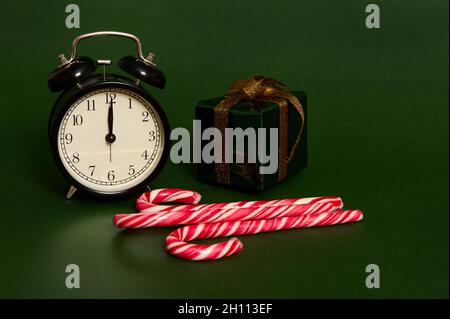 Nahaufnahme von schwarzem Wecker mit Mitternacht auf dem Zifferblatt, süßen gestreiften Lutschern, Süßigkeiten und Weihnachtsgeschenk in Glitter-Geschenkpapier und Gold Stockfoto