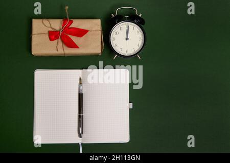 Flache Lay-Komposition mit einem Stift auf einem geöffneten Notizblock mit leeren leeren leeren Blättern, Wecker und einem Geschenk in Bastelpapier mit Schleife und rot gebunden Stockfoto