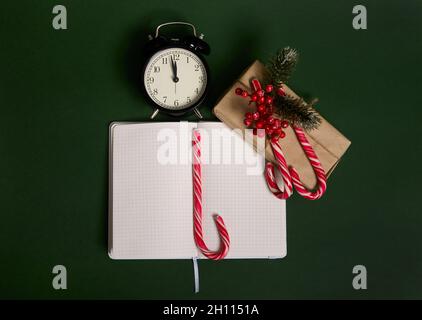 Hochwinkelansicht des Weckers mit Mitternacht, Weihnachtsgeschenk in Geschenkpapier, Stechpalme, Zuckerstöcke und Notizblock mit geöffneter Seite verpackt Stockfoto