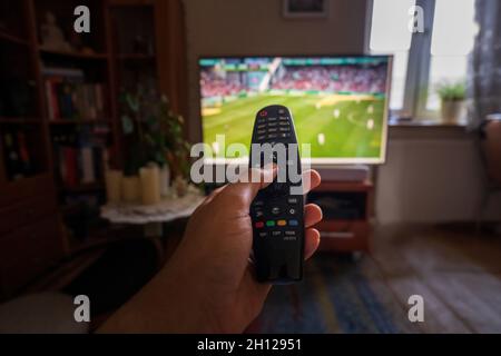 Eine handzeigende Fernbedienung zum fernseher oder Fernseher mit Fußball- oder Fußballspiel in Innenräumen, Blickwinkel, Weitwinkel mit selektivem Fokus. Stockfoto