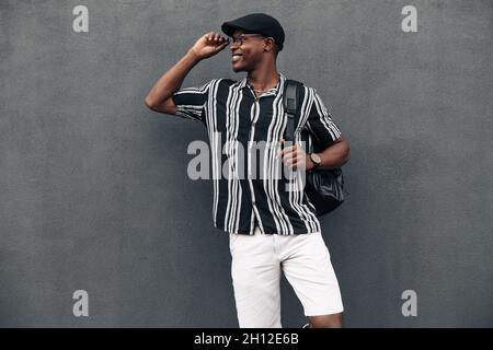 Porträt eines afroamerikanischen Mannes an einer schwarzen Wand, der zur Seite schaut, breit lächelt, ein Hemd trägt und eine schroppe Sommerstimmung hat Stockfoto