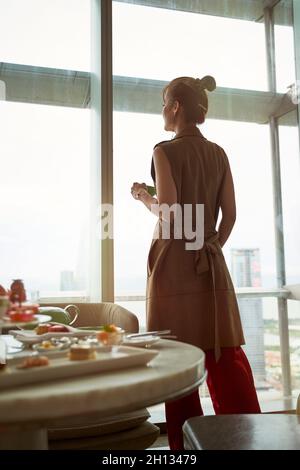 Reife asiatische Frau, die durch das Fenster des Hotelzimmers mit Frühstück auf dem Tisch die Aussicht betrat Stockfoto