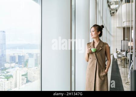 Reife asiatische Frau, die am Fenster steht und durch das Fenster schaut und eine Tasse Kaffee oder Tee hält Stockfoto