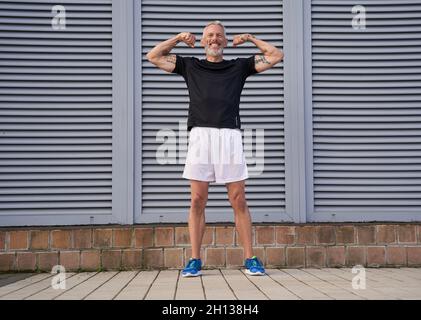 Ganzkörperaufnahme eines glücklichen tätowierten Mannes mittleren Alters in guter Form, lächelnd, während er seine muskulösen Arme, Bizeps und vor grauem Hintergrund im Freien posiert Stockfoto
