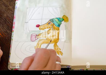 Hand mit Pinsel und unvollendete Aquarell Zeichnung des Hundes. Eine Frau färbt eine Bleistiftskizze auf Papier. Öffnen Sie das Notizbuch auf einem braunen Holztisch. Teil eines Stockfoto
