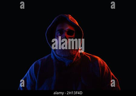 Mann mit Bart und Halbgesicht mit Knochen und Totenkopf in dunklem Hoodie, beleuchtet mit roten und blauen Lichtern Stockfoto