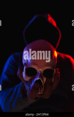 Dunkle Figur im Kapuzenpullover mit menschlichem Schädel in der Hand vorne, beleuchtet mit rotem und blauem Licht, Halloween-Konzept Stockfoto