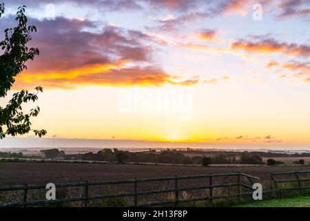 Der Sonnenaufgang über der Landschaft von Kent in England. Gepflügtes Feld und ein traditioneller Holzzaun im Vordergrund und darüber hinaus eine neblig bedeckte ländliche Ackerlandlandschaft, die zum Horizont führt. Dünnes Wolkenband, über dem die Sonne in einem gelben bis mauvigen Himmel mit einigen Wolken auftaucht. Stockfoto