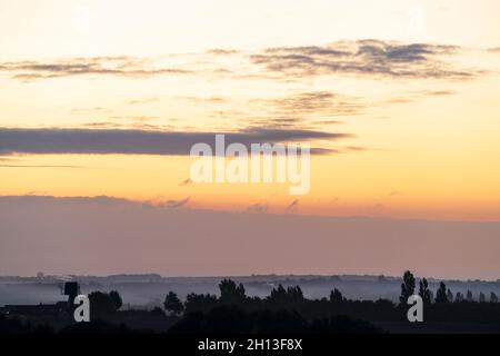 Der Morgenhimmel über der Landschaft von Kent in England. Eine einschiffige Windmühle, ohne die Segel, silhouetted gegen den Nebel, der zwischen Reihen von Bäumen aufsteigt, mit der Stadt Ramsgate Ion der Horizont weit in der Ferne. Gelb-orangefarbener Himmel mit einer dicken Wolkenschicht am Horizont und einigen niedrigen Cumuluswolken am offenen Himmel darüber. Stockfoto