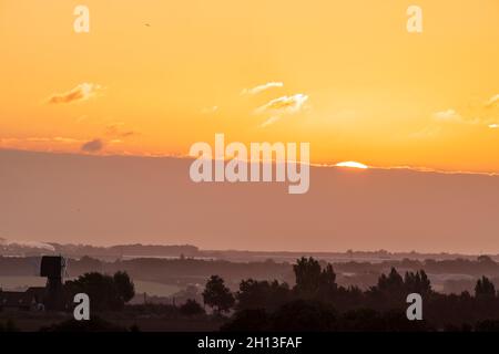 Der Sonnenaufgang über der Landschaft von Kent in England. Eine einschiffige Windmühle, ohne die Segel, silhouetted gegen den Nebel, der zwischen Reihen von Bäumen aufsteigt, mit der Stadt Ramsgate Ion der Horizont weit in der Ferne. Gelb-orangefarbener Himmel mit einer dicken Wolkenschicht am Horizont und einigen niedrigen Cumuluswolken am offenen Himmel darüber. Stockfoto