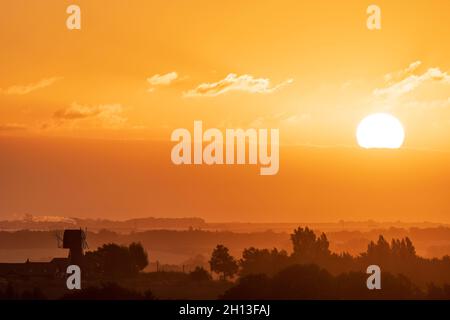 Der Sonnenaufgang über der Landschaft von Kent in England. Eine einschiffige Windmühle, ohne die Segel, silhouetted gegen den Nebel, der zwischen Reihen von Bäumen aufsteigt, mit der Stadt Ramsgate Ion der Horizont weit in der Ferne. Gelb-orangefarbener Himmel mit einer dicken Wolkenschicht am Horizont und einigen niedrigen Cumuluswolken am offenen Himmel darüber. Stockfoto