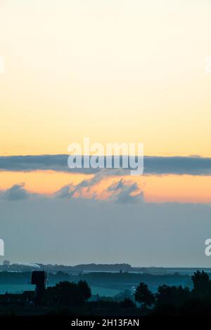 Der Morgenhimmel über der Landschaft von Kent in England. Eine einschiffige Windmühle, ohne die Segel, silhouetted gegen den Nebel, der zwischen Reihen von Bäumen aufsteigt, mit der Stadt Ramsgate Ion der Horizont weit in der Ferne. Gelb-orangefarbener Himmel mit einer dicken Wolkenschicht am Horizont und einigen niedrigen Cumuluswolken am offenen Himmel darüber. Stockfoto