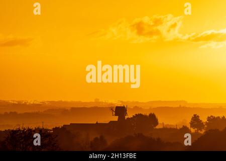 Der Sonnenaufgangshimmel über der Landschaft von Kent in England. Eine einschiffige Windmühle, ohne die Segel, silhouetted gegen den Nebel, der zwischen Reihen von Bäumen aufsteigt, mit der Stadt Ramsgate Ion der Horizont weit in der Ferne. Gelb-orangefarbener Himmel mit einer dicken Wolkenschicht am Horizont und einigen niedrigen Cumuluswolken am offenen Himmel darüber. Stockfoto